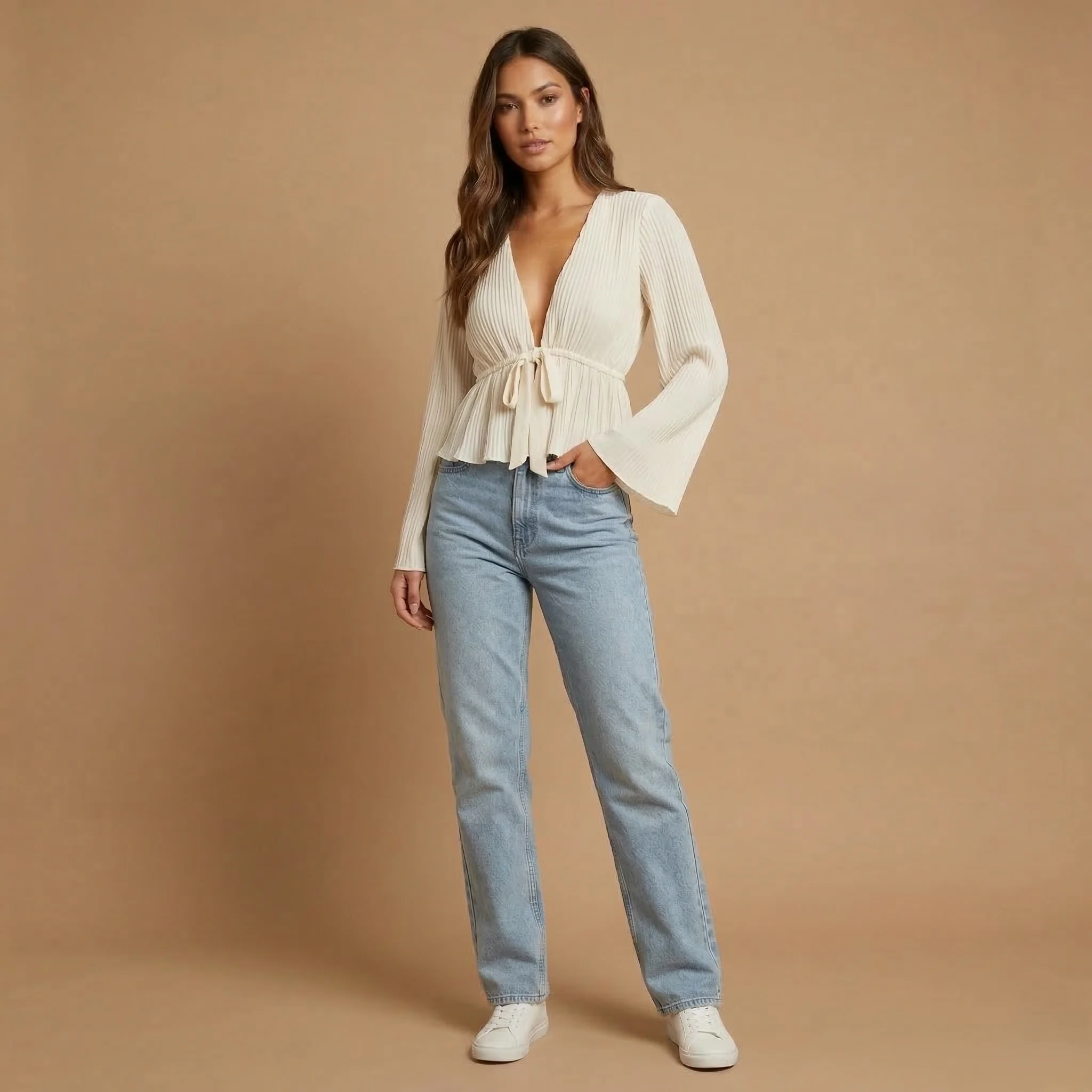 Woman wearing a white pleated peplum top with a deep V-neck, front tie detail, and bell sleeves paired with light blue jeans and white sneakers.